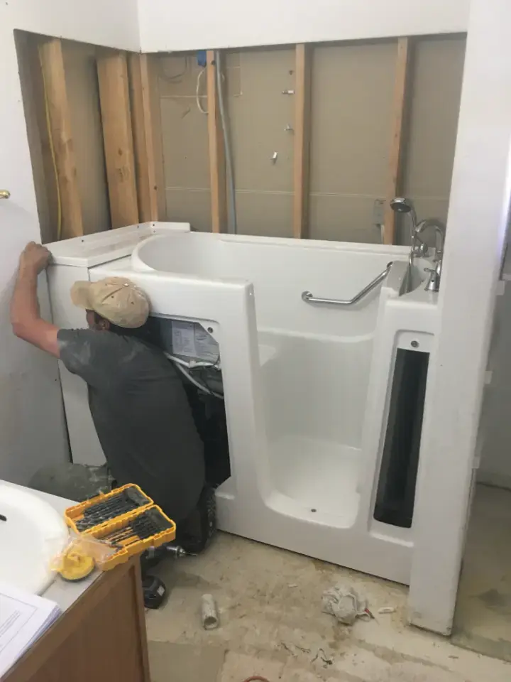 An installer setting a walk-in tub with a built-in seat and grab bar into an opened tub alcove